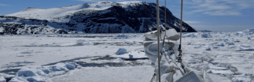 The bow of a boat as it cuts through water covered in snowy ice. Rocky land looms in the distance.